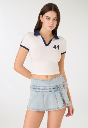 Biała krótka koszulka polo z granatowym kołnierzem i numerem 44, zestawiona z jasnoniebieską plisowaną mini spódniczką dżinsową z ozdobnymi klamrami.