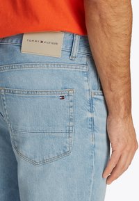 Blugi din denim albastru deschis, cu o etichetă din piele inscripționată "TOMMY HILFIGER" pe talie, având un mic accent cu logo pe buzunarul din spate.