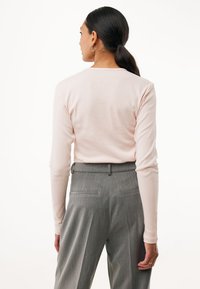 Frau mit dunklem Haar, das zu einem Pferdeschwanz gebunden ist, trägt ein hellrosa, geripptes Langarmoberteil und hochgeschnittene graue Hosen, von hinten zu sehen.