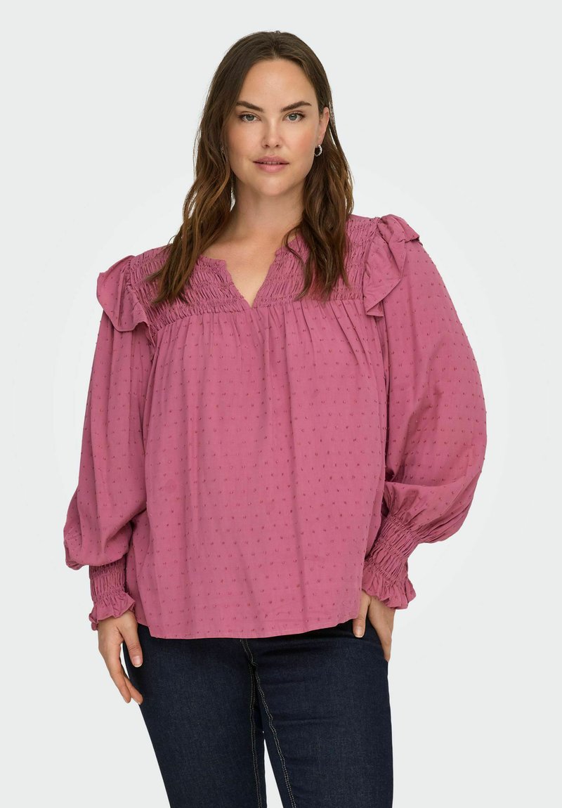 Kobieta z długimi brązowymi włosami nosi luźną różową bluzkę z falbaniastymi ramionami i mankietami, w połączeniu z ciemnoniebieskimi dżinsami.