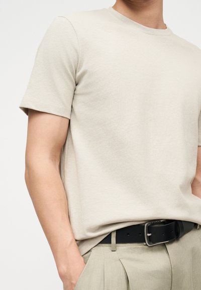 Άνδρας που φοράει μπεζ κοντομάνικη μπλούζα, κλεισμένη μέσα σε μπεζ πλισέ παντελόνι με μαύρη δερμάτινη ζώνη.