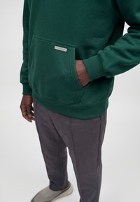 Homme portant un sweat à capuche vert foncé avec la main dans la poche avant, pantalon gris et chaussures beiges sur fond blanc.