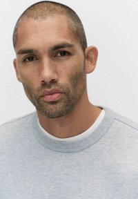 Harmaa verkkari, jossa on pyöreä kaula-aukko ja tekstuuriset yksityiskohdat, päällä valkoinen T-paita; tausta on yksinkertainen valkoinen.