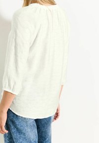 Blusa blanca texturizada con mangas tres cuartos, escote fruncido y un sutil detalle de costura, combinada con jeans de denim azul.