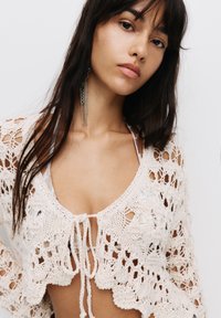 Cardigan croșetat în alb murdar, cu un design din plasă și margine ondulată, având un închizător tip cravată în față și o textură distinctivă.