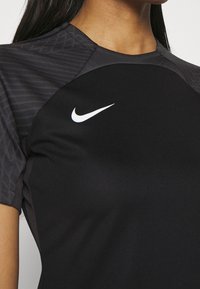 Top deportivo negro con un panel en el hombro de un gris oscuro texturizado y un logo de Nike blanco en el pecho, fabricado con un material transpirable.