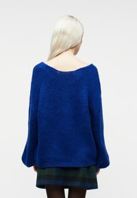 Kobieta z długimi blond włosami nosi przejrzysty niebieski sweter i spódnicę w kratę, zwrócona plecami na jednolitym białym tle.