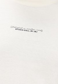 Baltas audinys su mažais juodais užrašais "Įkurtas devyniasdešimtųjų STUDIJA COLL. AUTORIUS J&J" sutelktas arti viršaus.