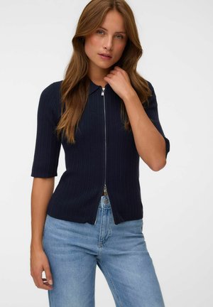 Woman with long brown hair wearing a zipped navy blue ribbed short-sleeve top and light blue jeans against a plain background.