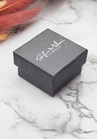 Svart texturerad smyckesask med ett lock, prydd med vit logotext. Asken har en kvadratisk form och en slät yta.