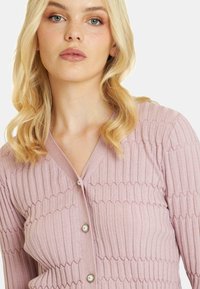 Jasnoróżowy kardigan w prążki z dekoltem w serek, z zapięciem na perłowe guziki oraz teksturowanym, falistym wzorem na tkaninie.