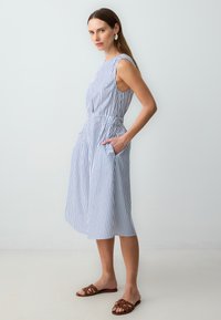 Gestreiftes ärmelloses Kleid aus hellblauem und weißem Stoff, in der Taille gerafft mit seitlichen Taschen. Getragen mit braunen flachen Sandalen.
