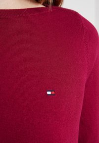 Gros plan d’une personne portant un pull en maille rouge avec un petit logo rectangulaire rouge, blanc et bleu marine sur la poitrine.