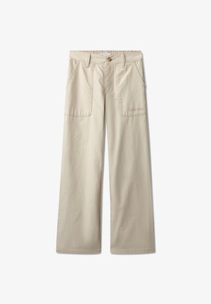 Junge steht mit den Händen in den Taschen, trägt ein weißes Calvin Klein T-Shirt, beige weit geschnittene Hose und weiße Turnschuhe vor hellgrauem Hintergrund.