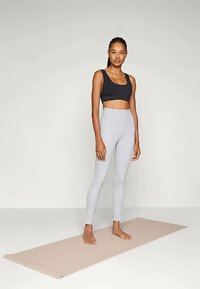 Kvinna som står barfota på en beige yogamatta, iklädd en svart sport-bh och ljusgrå högmidjade leggings mot en enfärgad bakgrund.