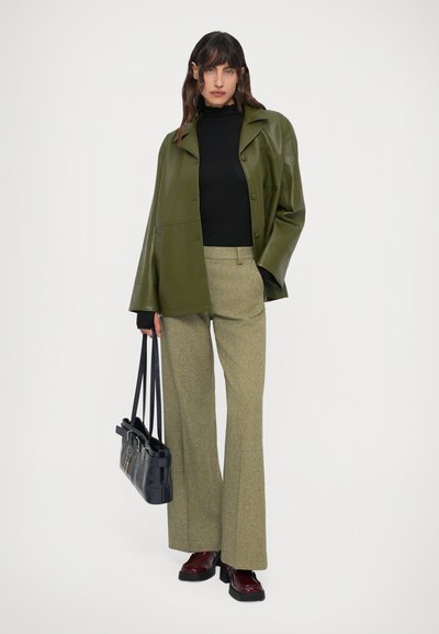 Giacca in ecopelle verde sopra una maglietta nera, abbinata a pantaloni verdi chiari texturizzati e scarpe col tacco bordeaux. Tiene una borsa nera.