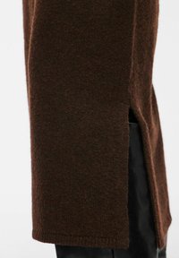 Gros plan d'un tissu tricoté brun foncé avec une fente sur la couture latérale, révélant un vêtement noir en dessous.