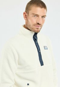 Pull en polaire blanc cassé avec un col haut, patte de boutonnage bleu marine et un patch logo de montagne sur la poitrine. Détail de tissu texturé.