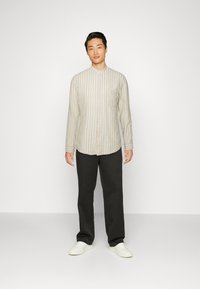 Camicia a righe beige a maniche lunghe con bottone frontale e taschino sul petto, abbinata a pantaloni scuri e sneakers bianche, in piedi contro uno sfondo neutro.