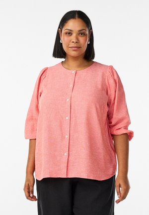 Kvinde med skulderlangt sort hår iført en koralfarvet knap-op-bluse og sorte bukser, stående mod en hvid baggrund.