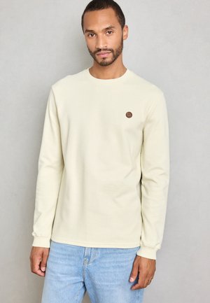 Hombre con pelo corto y barba, vestido con una camisa de manga larga color crema con un pequeño logo marrón en el pecho y jeans de color azul claro, de pie contra una pared gris.