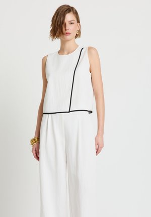 Jeune femme portant un haut blanc sans manches avec un liseré noir et un pantalon blanc ample, accessoirisée avec des bracelets et des boucles d'oreilles dorés.