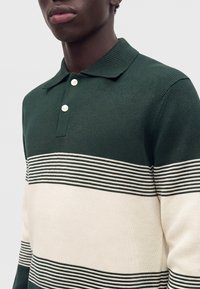 Homme portant un polo en maille vert foncé et crème avec des rayures horizontales et deux boutons blancs sur le col.