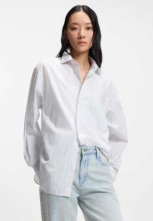 Young woman with long dark hair wearing a white striped button-up shirt tucked into light blue jeans, standing against a plain background.