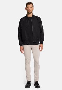 Zwarte bomberjack met een rits aan de voorkant en een zijzak, gecombineerd met lichtgekleurde slim fit broek en zwarte veterschoenen.