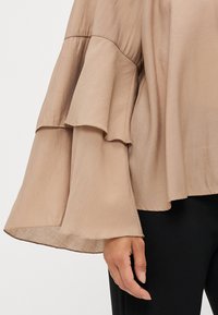 Light beige blouse with ruffled bell sleeves, made of soft, flowing fabric. Smooth texture and layered hem detail. Worn over black pants.