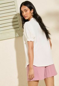 Top bianco con maniche a sbuffo e polsini arricciati abbinato a pantaloni corti rosa chiaro. Tessuto liscio, vista posteriore che mostra la vestibilità comoda e lo stile informale.