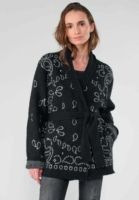 Cardigan en tricot noir avec des motifs décoratifs blancs, ouverture à l'avant, ceinture à nouer à la taille et manches longues. Tissu texturé avec des poignets contrastés.