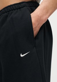 Χέρι που ξεκουράζεται μέσα στην τσέπη μαύρου παντελονιού Nike με ελαστική μέση και λευκό κεντημένο λογότυπο.