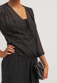 Blusa negra con detalle de lazo, tela texturizada y líneas verticales plateadas. Combinada con pantalones de cintura alta y un pequeño bolso de mano negro.