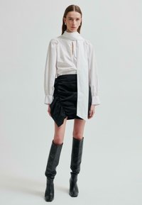 Žena v bílé blůze s dlouhým rukávem a detailem šály, černé skládací mini sukni a černých kožených vysokých botách stojí proti jednobarevnému pozadí.