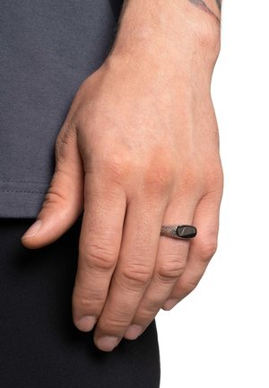 Anello in metallo con una fascia di argento texturizzata e una pietra nera ovale incastonata, indossato su una mano su uno sfondo grigio.