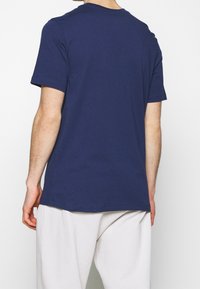 Henkilö nähtynä takaa, joka käyttää yksinkertaista tummansinistä lyhythihaista T-paitaa ja löysiä valkoisia housuja valkoista taustaa vasten.