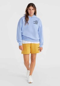 Hele sinine kapuutsiga särk, millel on must trükk "The Future Surf Society", kombineeritud kollaste lühikesed püksid, millel on sarnane logo, ja valged tossud.