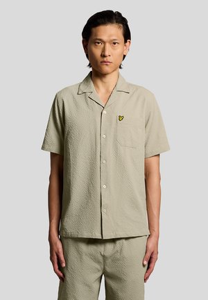 Mann trägt ein beige strukturiertes kurzärmeliges Hemd mit Knöpfen und passende Shorts, steht vor einem schlichten hellgrauen Hintergrund.