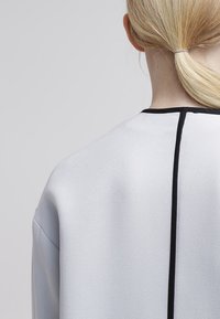 Vaaleanharmaa paita, jossa on sileä pinta ja musta reunus kaula-aukolla sekä keskisauma takana. Muotoilu on minimalisti.