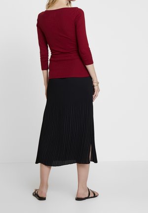 Vrouw die een nauwsluitend donkerrood geribbeld shirt draagt met driekwart mouwen en een zwarte geplooide midi-rok met een zijspalk, staande op blote voeten.