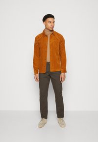 Brown corduroy shirt jacket with button closure, paired with a beige turtleneck and dark green trousers. Shoes are light beige suede.
