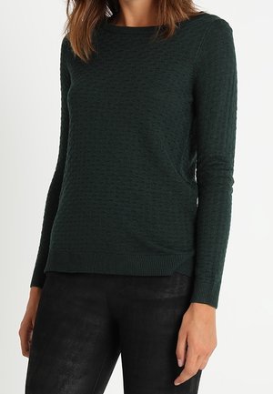 Femme portant un pull à manches longues texturé vert foncé associé à un pantalon noir ajusté, debout devant un fond clair uni.