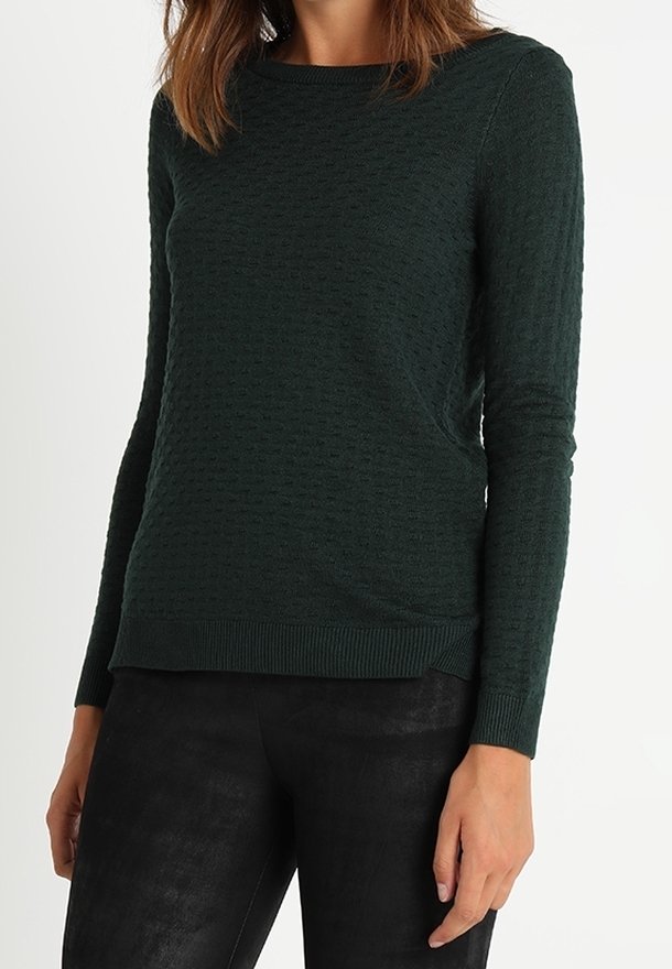 Femme portant un pull à manches longues texturé vert foncé associé à un pantalon noir ajusté, debout devant un fond clair uni.