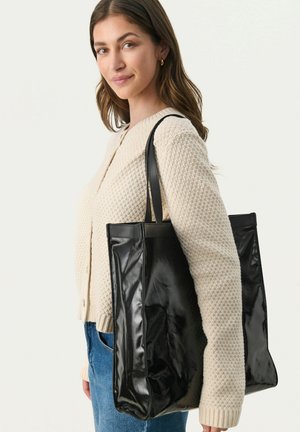 Schwarze Leder-Tote-Bag mit glatter Textur, rechteckiger Form und doppelten Schulterriemen. Modell trägt einen hellbeigen Strickpullover.