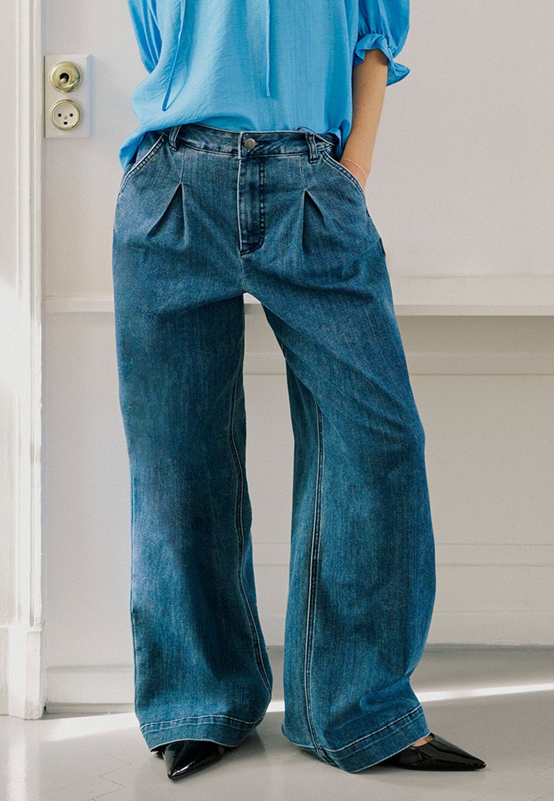 Pantalon en jean bleu à jambes larges avec des plis à l'avant, présentant des surpiqûres contrastantes et une taille haute, associé à des talons noirs pointus.