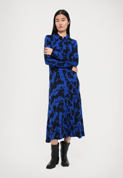 Haljina plavo-crnog uzorka s dugim rukavima, ovratnikom i širokom suknjom. Model nosi crne gležnjače. Glatka tekstura.