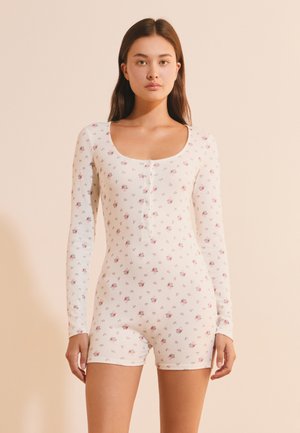 Junge Frau mit langen dunklen Haaren, die einen weißen Langarm-Overall mit kleinen rosa Blumenmustern trägt, stehend vor einem beigen Hintergrund.