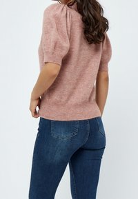 Žena v růžovém svetru s krátkým rukávem a tmavě modrých džínech, zobrazená zezadu na světlém jednobodovém pozadí.