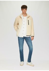 Beige fleecejack met een hoge kraag en een ritsborstas, gecombineerd met een wit shirt en blauwe distressed jeans. Witte sneakers maken de look af.